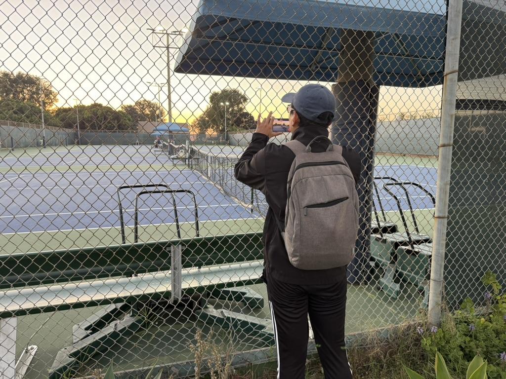A tennis parent filming a court through a chain-link fence at sunset